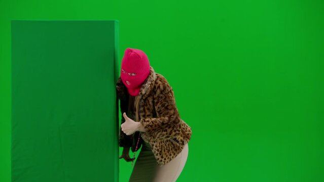 A woman in pink balaclava, tiger coat and dress jumps out from behind a green banner, points her finger at it and shows a thumbs up gesture. Freak woman on green background in studio.