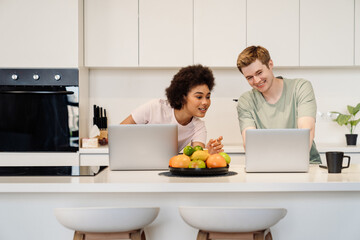 Cheerful couple using laptops while working together remotely sitting in kitchen