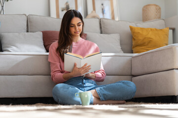 Pretty young woman reading a book and drinking a cup of coffee while sitting on the floor at home.