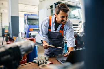 Happy mechanic using laptop while working at truck repair workshop.
