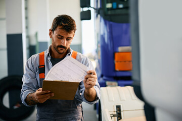 Truck maintenance mechanic going through notes while examining vehicle in workshop.