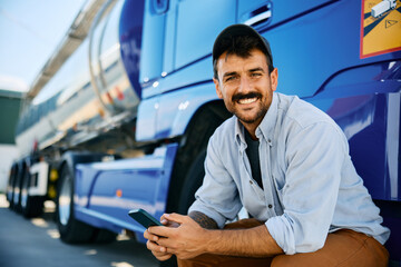 Happy truck driver using cell phone on parking lot and looking at camera. - obrazy, fototapety, plakaty