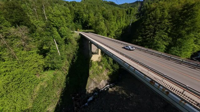 Aerial view luxury white sport car riding on bridge serpentine asphalt road at summer sunny greenery forest landscape. FPV sports drone flight over speed driving automobile at alpine resort scenery 4k
