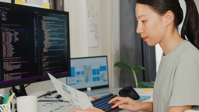 Side view of professional young Asia girl IT development programmer typing on keyboard coding programming fixing data code on computer screen and laptop on table in workroom at house office.