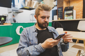 Bearded male using smartphone at modern coffee shop and chatting online messaging on mobile phone. Social networks and reading blog concept