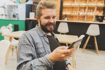 Bearded male using smartphone at modern coffee shop and chatting online messaging on mobile phone. Social networks and reading blog concept