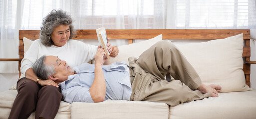 Asian grandfather lying on grandmother on couch and reading book in the living room. Retired couple...