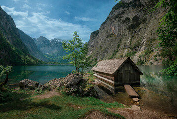 Fototapeta na wymiar Boathouse with mountain cliffs on lake Obersee at Berchtesgaden Bavaria, clouds in blue sky, turquoise calm water, Berchtesgaden Bavaria.