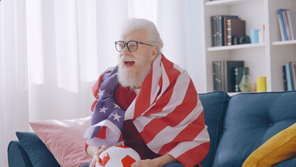 A man in his 60s, wrapped in the US flag, watches a soccer game, cheering enthusiastically for his...