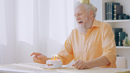 A gray-haired man sits in a birthday hat, about to blow out the candles on a cake, celebrating his...