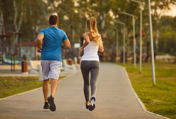 Energize your morning. Active young sports couple doing sports running together in city park in morning. Rear view of slim woman and man in sportswear jogging together in park on jogging track. - obrazy, fototapety, plakaty