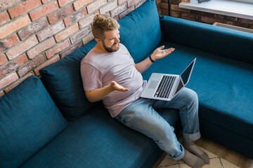 Millennial man at home on couch talking by video call with a laptop. Technologies for freelancer and communication online concept