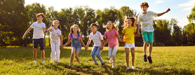 Teenage happy kids friends jumping on green grass in the summer park standing in a line. Smiling children boys and girls having fun together outdoors on a sunny day in nature. Banner.