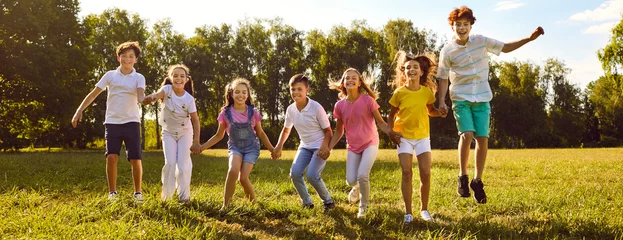 Fotobehang Teenage happy kids friends jumping on green grass in the summer park standing in a line. Smiling children boys and girls having fun together outdoors on a sunny day in nature. Banner. © Studio Romantic