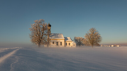 Bavarian church of Raisting with trees and snow and mist during winter and sunset, snow field in...