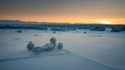 Bavarian church of Raisting with trees and snow during winter and sunset from above, snow field in the foreground, Bavaria Germany. - 702369904
