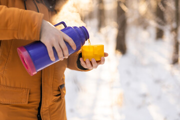 Man pours hot tea from a thermos into a snow walking in snowy frozen winter forest at sunset....