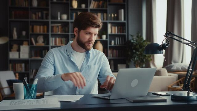 young focused man with beard in blue shirt sits at desktop in home office, works at laptop computer. takes document papers, look through thoughtfully. types on keyboard, smiles and nods