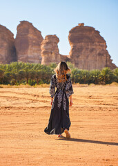 Young woman wearing long dark dress, walking on desert landscape, palm trees and large rock...