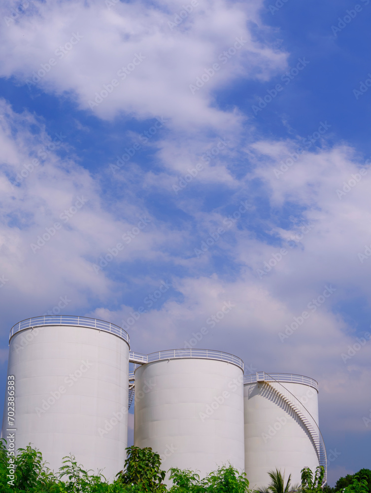 Wall mural three white storage fuel tanks behind row of green trees against white clouds on blue sky background