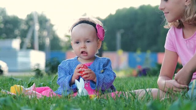 Cute baby girl with her sister in bunny ears playing with Easter eggs while resting on a grass outdoors 