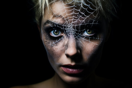 A woman with spiderweb makeup around one eye against a dark background.