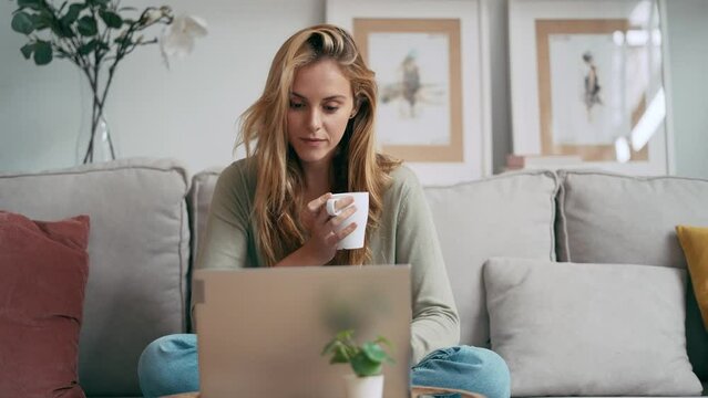 Video of beautiful young woman working with her laptop while drinking a cup of coffee sitting on a couch at home