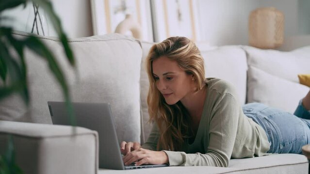 Video of pretty young woman working with her laptop while lying on couch at home