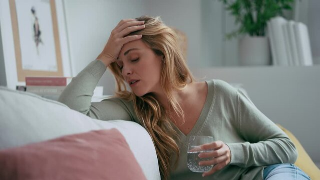 Video of unhealthy young woman with headache putting a pill in a glass of water sitting on sofa at home