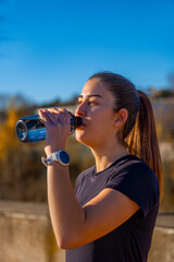 Beautiful young runner, beautifully illuminated by dawn sunlight, drinking and hydrating from a bottle of water, dressed in black sportswear, looking at the horizon in a park.