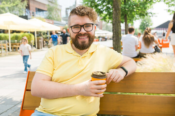 Millennial smiling student man in yellow polo t-shirt eyeglasses sitting on bench drink coffee have...