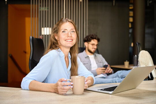 Businesswoman drinking tea or coffee near blurred colleague using smartphone