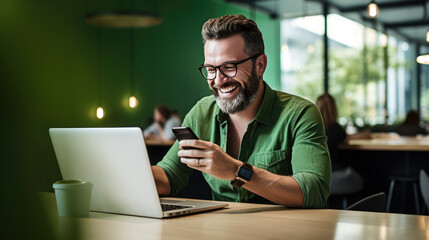 Cheerful man with a beard and glasses working on a laptop at a wooden desk