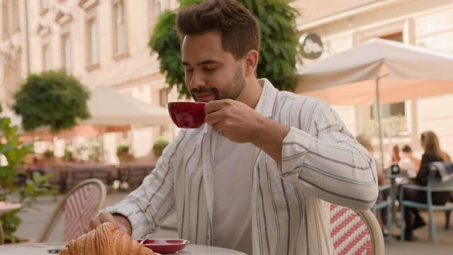 Young Caucasian man drinking delicious tasty hot coffee tea terrace cafe restaurant city street outside guy enjoying breakfast smiling aromatic businessman satisfied male relax morning with croissant