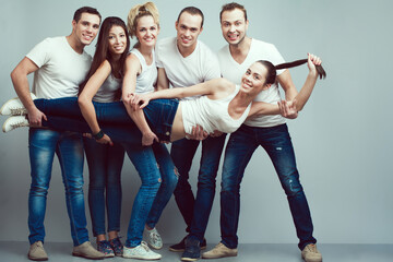 Obraz premium Happy together concept. Group portrait of healthy boys and girls in white t-shirts, sleeveless shirts and blue jeans holding friend in hands and posing over gray background. Urban style. Copy-space