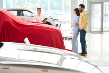 Car dealership consultant presents a car under a red cover
