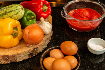 Bancada de mármore preta de cozinha com ingredientes como tigela de ovos inteiros, uma de molho de tomate, pote de sal e uma tábua de madeira com pimentões, abobrinha, cabeça de alho e cebola.