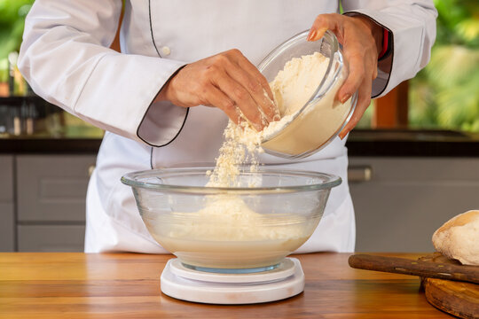 detalhe das m&atilde;os da chef de cozinha segurando em uma das m&atilde;os uma tigela de vidro com farinha de trigo e com a outra m&atilde;o jogando um punhado de trigo em outra tigela preparando a receita.