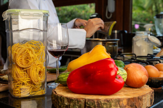Bancada de cozinha com piment&otilde;es amarelo e vermelho, cebola e abobrinha sobre uma t&aacute;bua de tronco, um pote de massa de talharim crua, uma ta&ccedil;a de vinho e chef de cozinhando ao fundo.