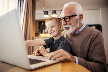 A cheerful grandfather and grandson sitting at home and smiling at the laptop.