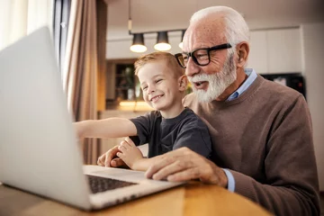Foto op Canvas A cheerful grandfather and grandson sitting at home and smiling at the laptop. © dusanpetkovic1