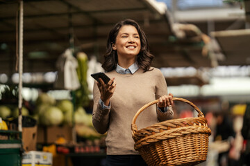 A woman with basket in hands is using a phone while shopping at local market.