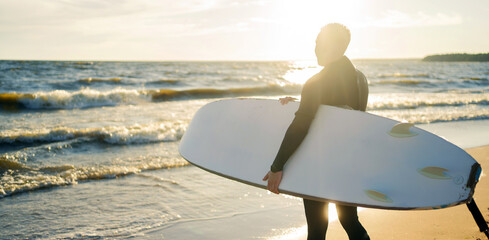 Male surfer in wetsuit with surfboard at sunset, ready for an evening session on the waves.