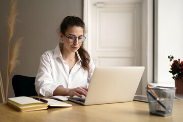 Concentrated young woman working on a laptop in a serene home office, displaying focus and...