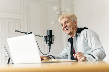 Happy podcaster recording in a bright room, speaking into a microphone with a laptop and notes.