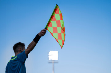 Raised flag of the sideline referee during football match, blue sky and lamps in the background.