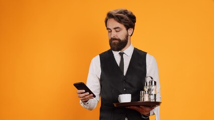 Catering employee texting messages on phone while carrying restaurant tray with drinks and cutlery,...