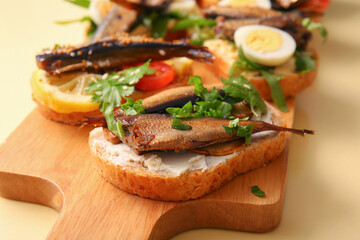 Wooden board of tasty sandwiches with canned smoked sprats on yellow background