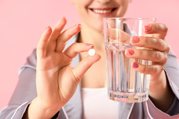Young woman in pajamas with pill and glass of water on pink background, closeup