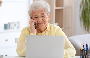 Fototapeta premium Senior woman with laptop talking by mobile phone at home